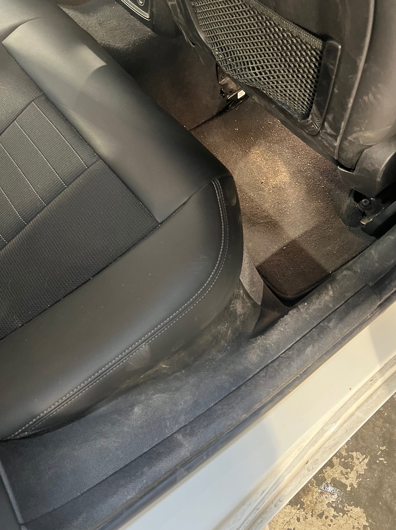 Wnętrze samochodu przed czyszczeniem: zabrudzona tapicerka i podłoga w okolicy tylnego siedzenia, widoczna siatka na oparciu fotela.