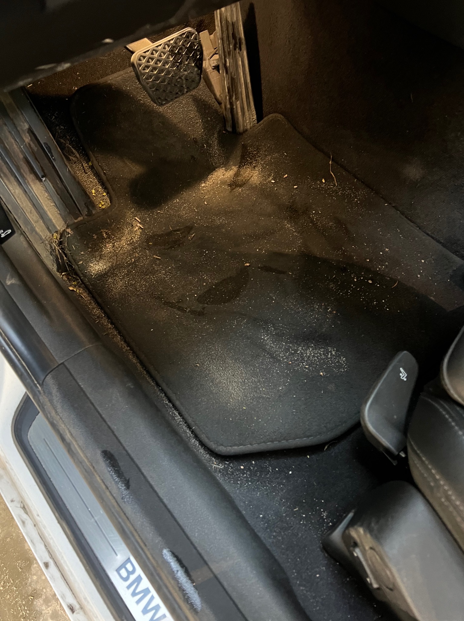 Wnętrze samochodu BMW przed czyszczeniem: brudny dywanik kierowcy z piaskiem, okruchami i resztkami roślin, widoczny pedał i fragment progu.