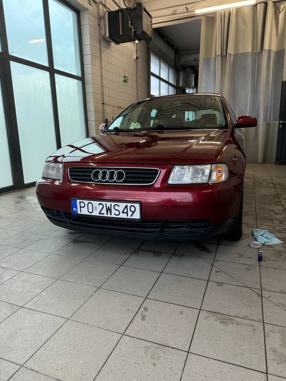 Czerwony samochód Audi A3 po umyciu w myjni, widoczna wilgotna powierzchnia i resztki wody na podłodze z płytek, w tle zasłona oddzielająca stanowiska.