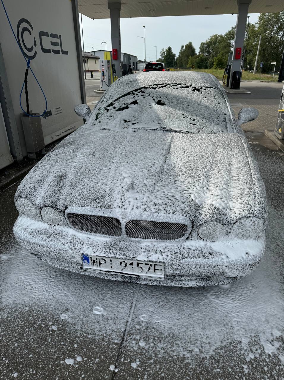 Samochód pokryty pianą podczas mycia na myjni samoobsługowej, widoczny numer rejestracyjny i elementy konstrukcyjne myjni.