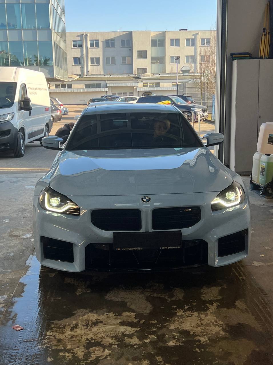 Białe BMW M2 stoi wewnątrz myjni, widoczne mokre ślady po umyciu na posadzce, w tle budynek biurowy i inne samochody.