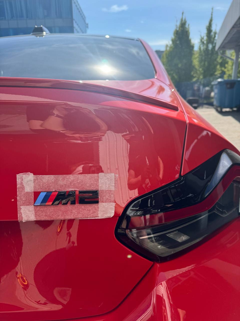 Czerwony samochód z nowym emblematem M2 przymocowanym taśmą na klapie bagażnika, w trakcie montażu.