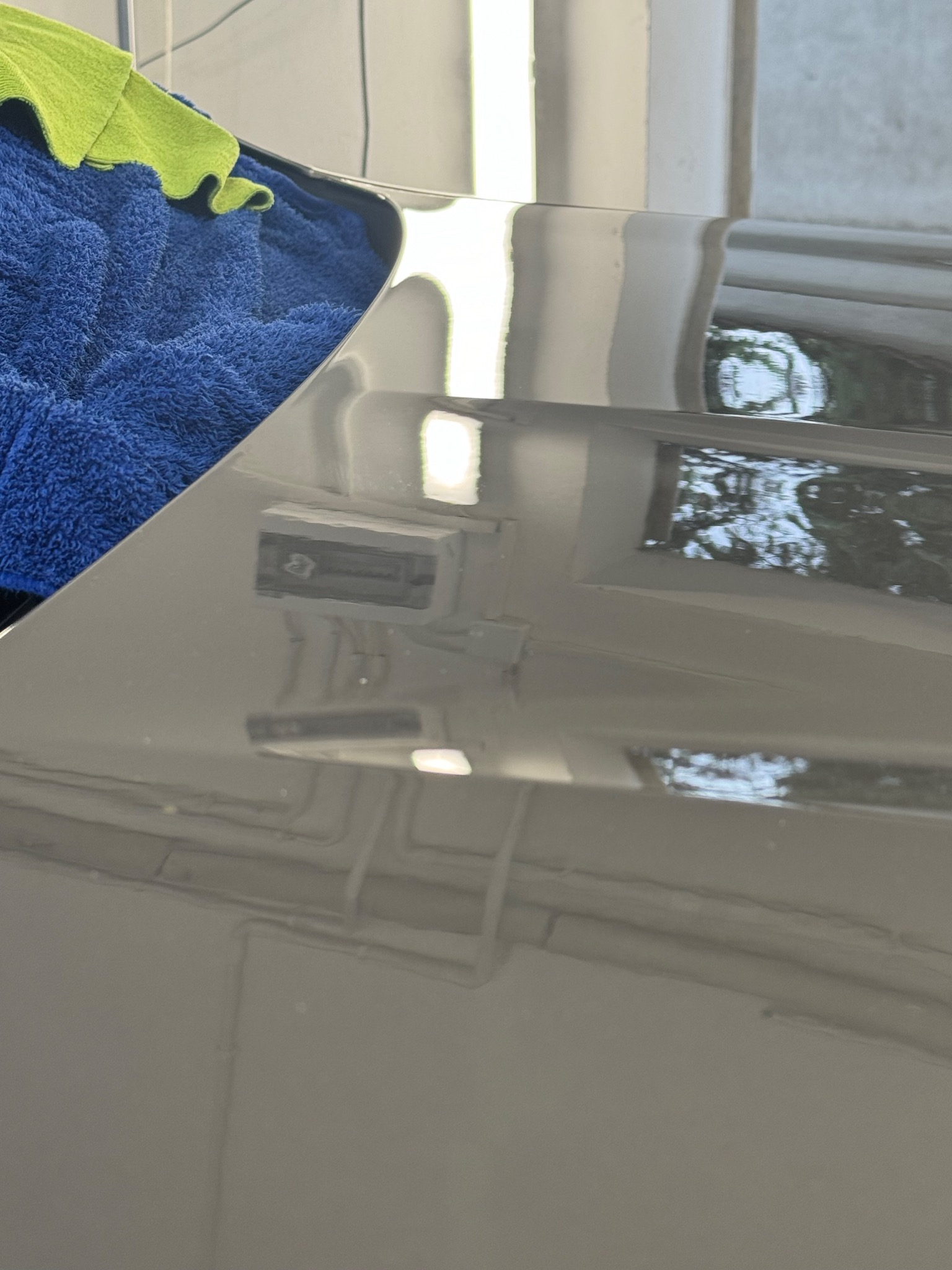 Błyszcząca, szara karoseria samochodu po umyciu, z widocznymi odbiciami otoczenia, obok leżą niebieska i zielona ściereczka z mikrofibry.