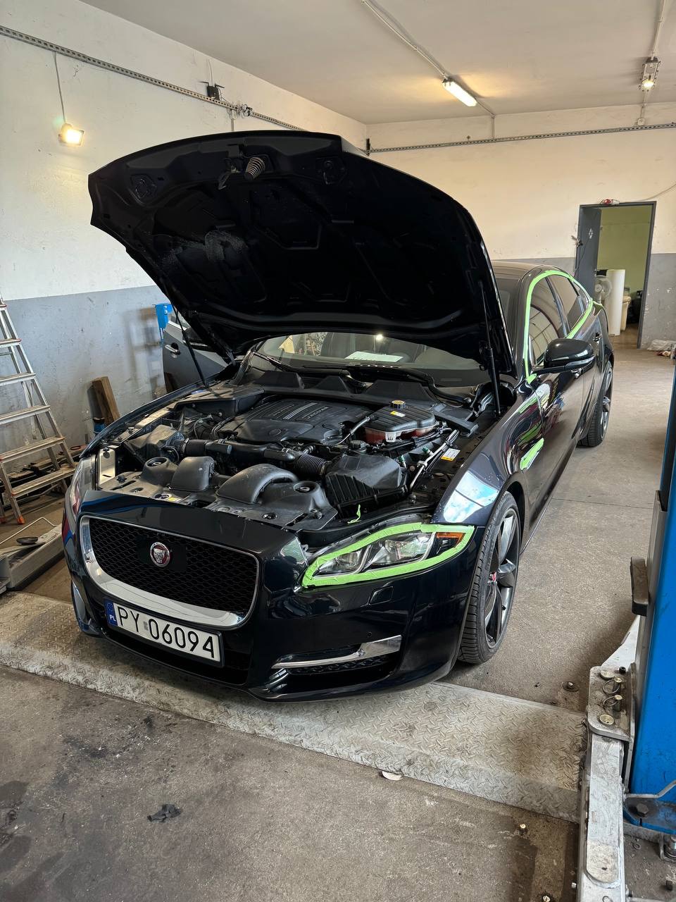 Czarny Jaguar XJ z otwartą maską, zabezpieczony taśmą malarską wokół reflektorów, silnik widoczny, na podnośniku w warsztacie.