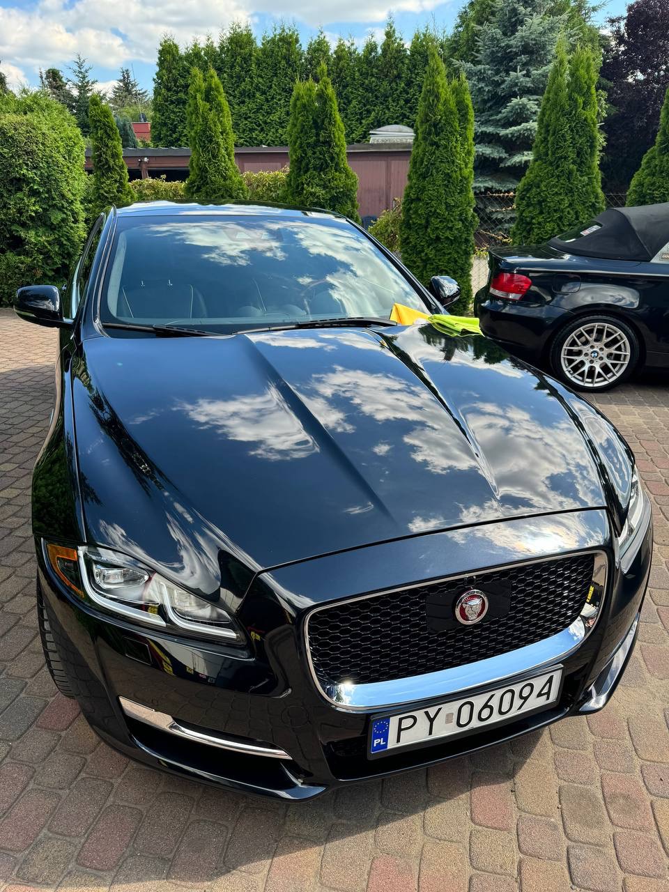 Błyszczący, czarny Jaguar XJ po umyciu, z widocznym odbiciem chmur na karoserii i żółtą ściereczką na masce, zaparkowany na kostce brukowej przed zadbanym ogrodem.