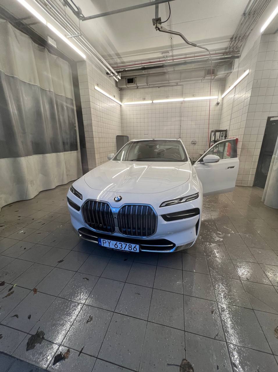 Białe BMW serii 7 po umyciu, z otwartymi drzwiami w jasnej myjni samochodowej z szarymi zasłonami i mokrą podłogą.