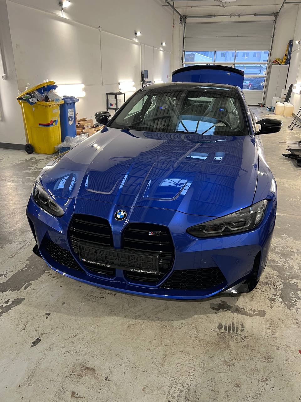 Błyszczący, niebieski samochód BMW M3 z otwartym bagażnikiem, z folią ochronną na siedzeniach, stojący w jasnym garażu z żółtym i niebieskim koszem na śmieci w tle.