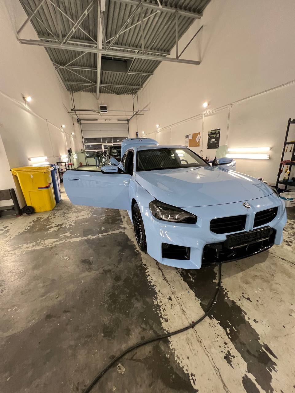 Jasnoniebieski samochód z otwartymi drzwiami w warsztacie, widoczny żółty kosz na śmieci i narzędzia na półce w tle.