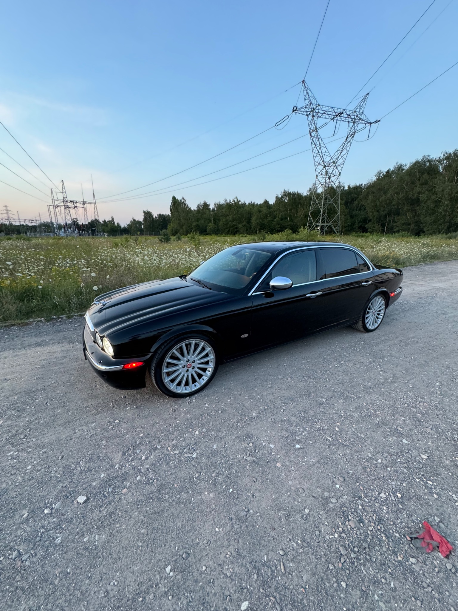 Czarny Jaguar XJ na szutrowej drodze z widokiem na pole z kwiatami i słupy wysokiego napięcia w tle.