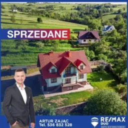 Zdjęcie domu z czerwoną dachówką na tle zielonych wzgórz, z nakładką 'SPRZEDANE' i wizerunkiem agenta nieruchomości oraz logo RE/MAX.
