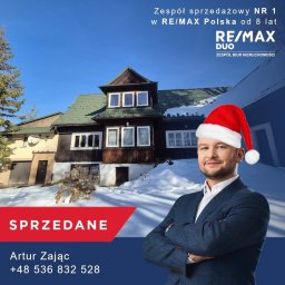Artur Zając - Agent nieruchomości w czapce Mikołaja na tle sprzedanego, zimowego domu w stylu góralskim. Logo RE/MAX DUO i napis SPRZEDANE.