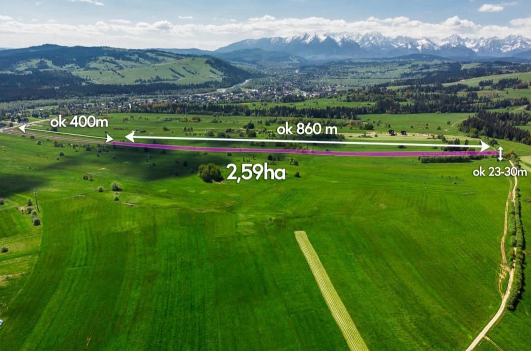 Zdjęcie z lotu ptaka przedstawia rozległą, zieloną działkę zarysowaną fioletową linią, z podanymi wymiarami: ok. 400m, ok. 860m, ok. 23-30m oraz powierzchnią 2.59ha; w tle widoczne pasmo górskie...