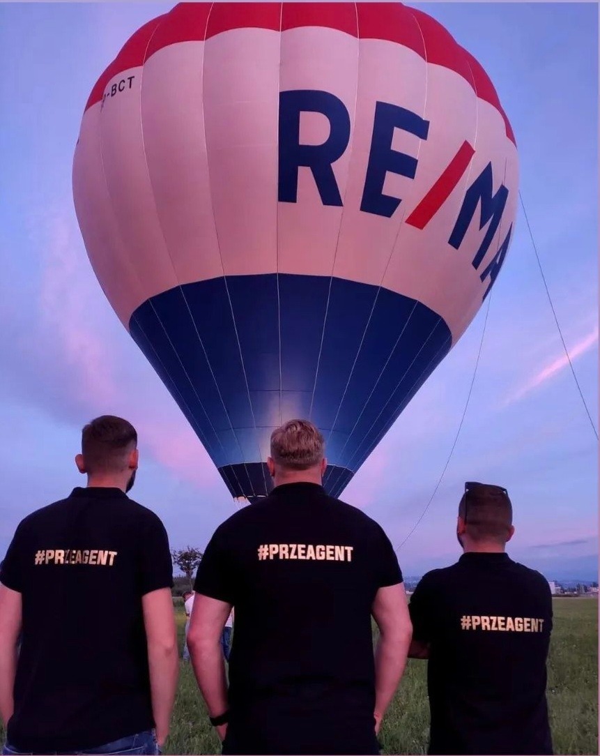 Trzech mężczyzn w czarnych koszulkach z napisem #PRZEAGENT stoi tyłem do aparatu, patrząc na balon z logo RE/MAX na tle zachodzącego słońca.