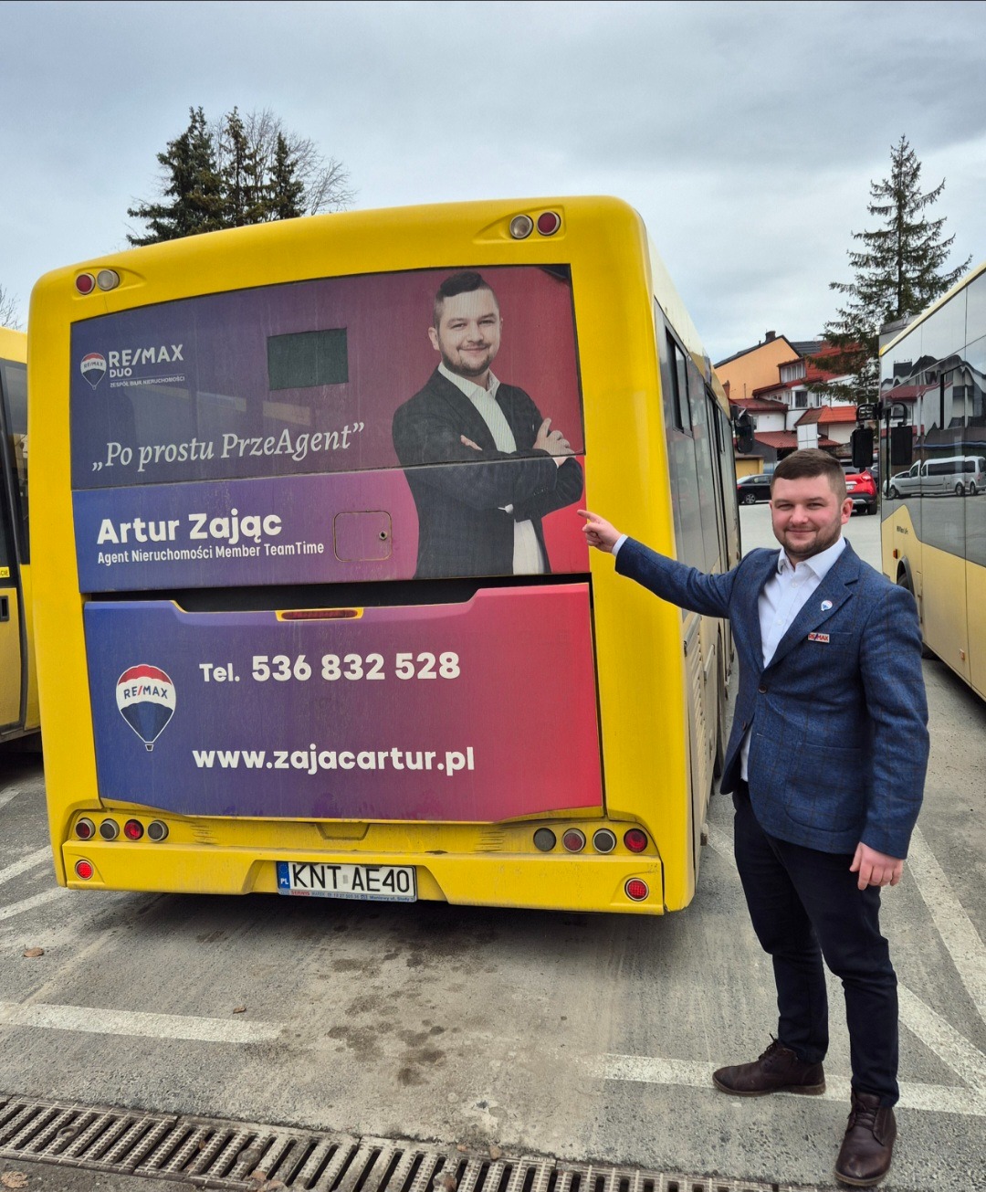 Mężczyzna w niebieskiej marynarce wskazuje na reklamę agenta nieruchomości na żółtym autobusie, widoczny numer telefonu i adres strony internetowej.