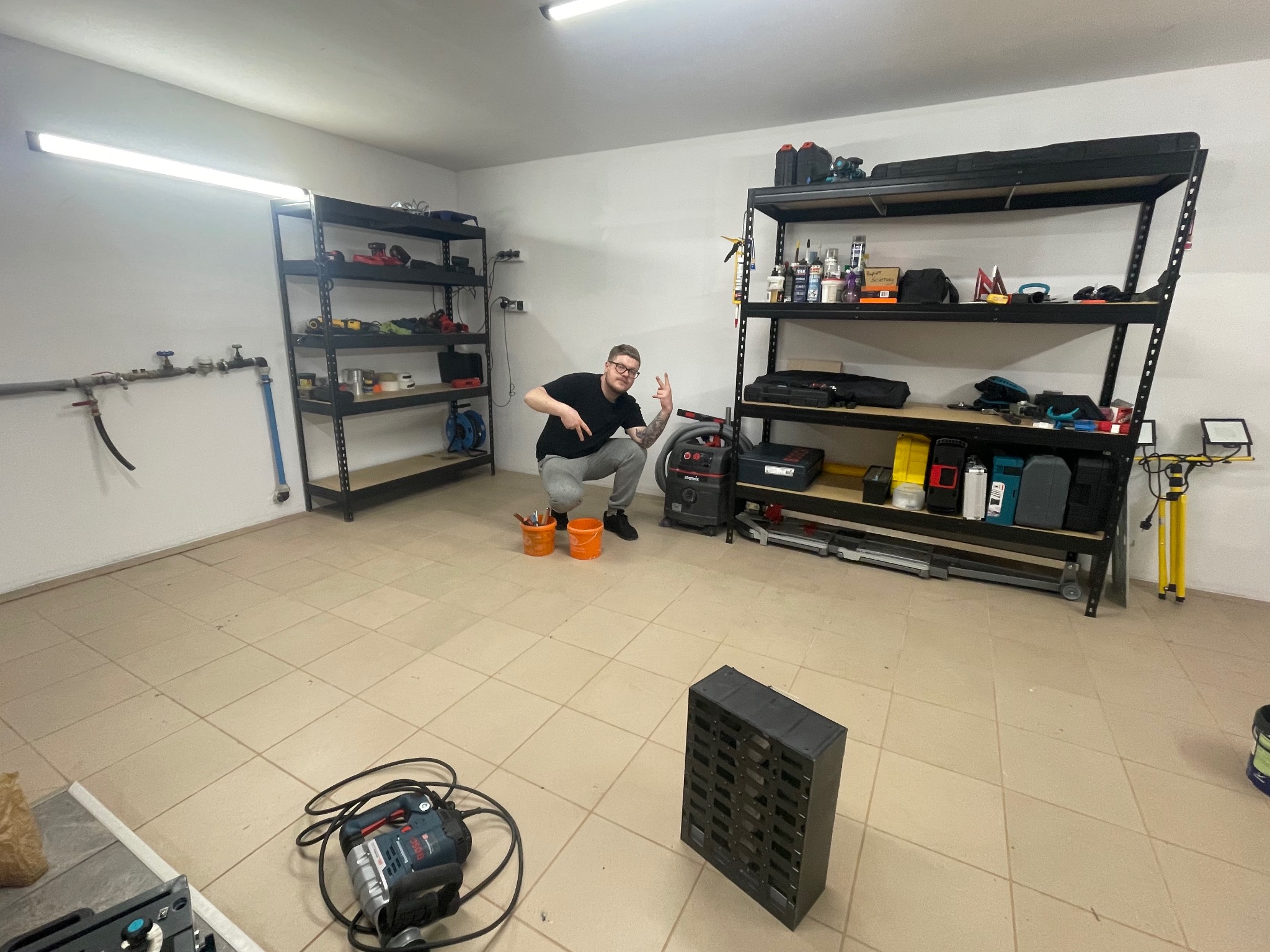 Mężczyzna kuca w garażu z narzędziami, prezentując gest ręką, obok odkurzacza przemysłowego i dwóch pomarańczowych wiader z narzędziami, z regałami wypełnionymi sprzętem w tle.