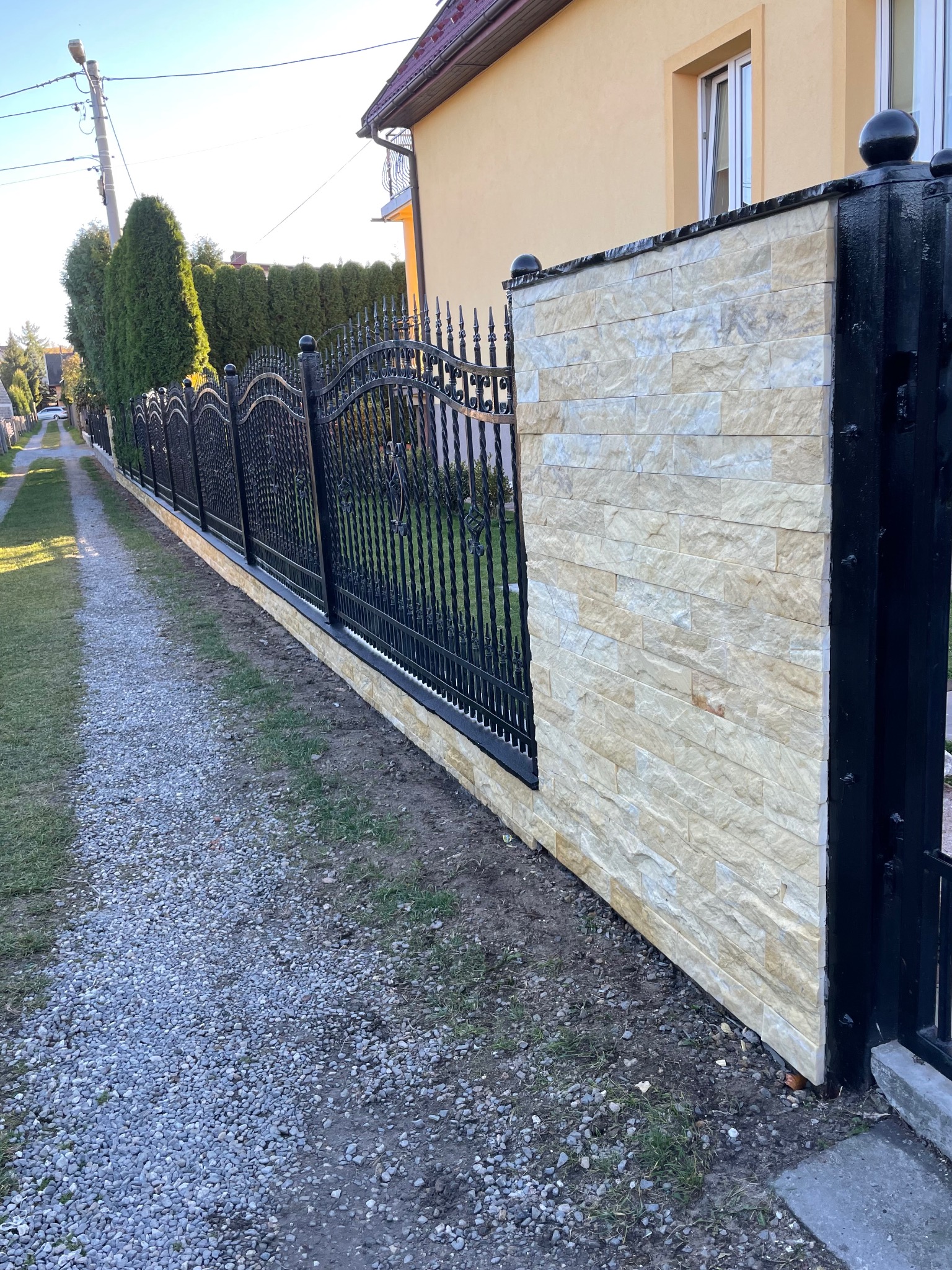 Nowoczesne ogrodzenie z czarnymi, metalowymi przęsłami i fragmentami wykończonymi jasnym kamieniem elewacyjnym, widok z boku na tle zieleni i ścieżki.