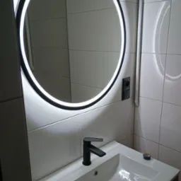 Nowoczesna łazienka z okrągłym lustrem LED, czarną armaturą i białą umywalką nablatową. Na blacie widoczny fragment niebieskiej taśmy malarskiej.