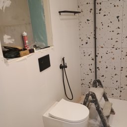 Jan -Bud - Remont łazienki w Strzyżowie: podwieszana toaleta, czarna armatura, płytki lastryko na ścianie, drabina malarska i narzędzia w trakcie montażu.