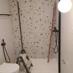 Jan -Bud - Nierównomiernie ułożone płytki lastryko na ścianie łazienki w trakcie prac wykończeniowych, widoczna drabina malarska, toaleta i czarna armatura prysznicowa.