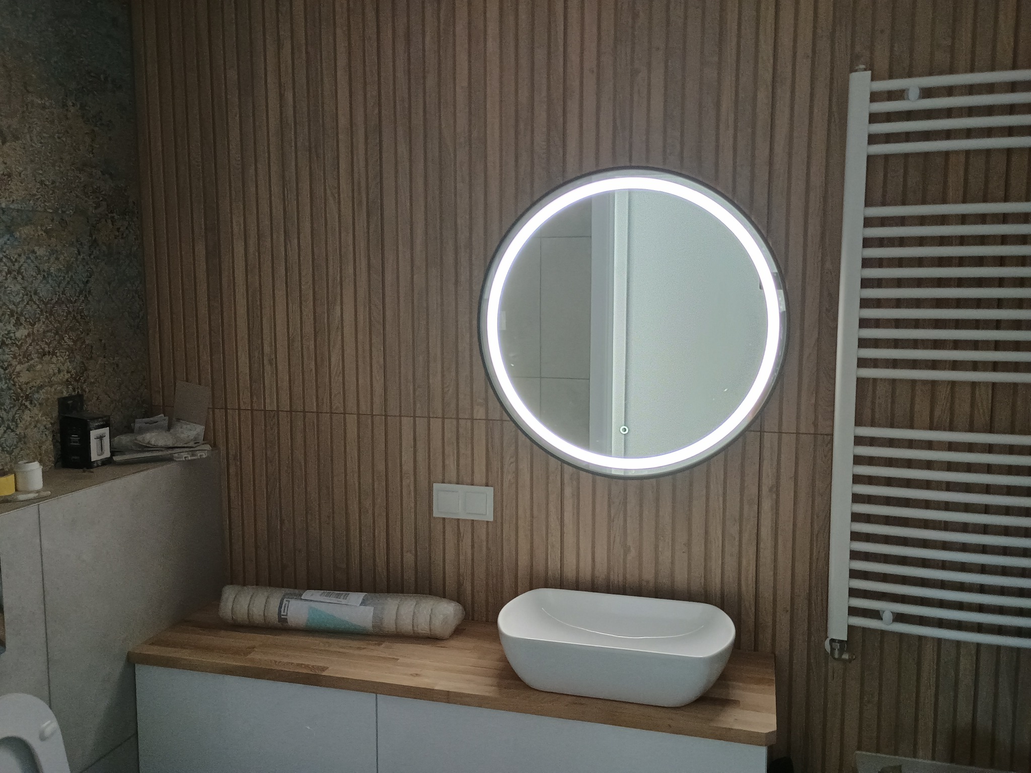 Nowoczesna łazienka z umywalką nablatową, okrągłym lustrem z podświetleniem LED i grzejnikiem drabinkowym na ścianie wyłożonej panelami drewnopodobnymi. Widoczny zwój tapety na blacie i fragment...