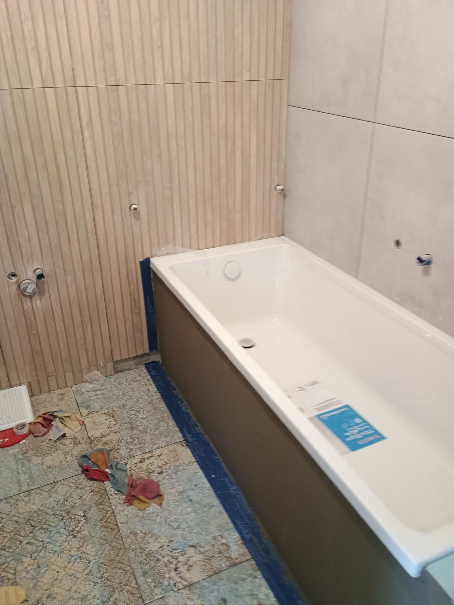 Nowa, biała wanna z szarym obiciem, zabezpieczona niebieską taśmą, stoi w łazience z drewnianą ścianą z pionowymi żłobieniami i szarą ścianą z płytek, na podłodze widoczne ślady prac remontowych...