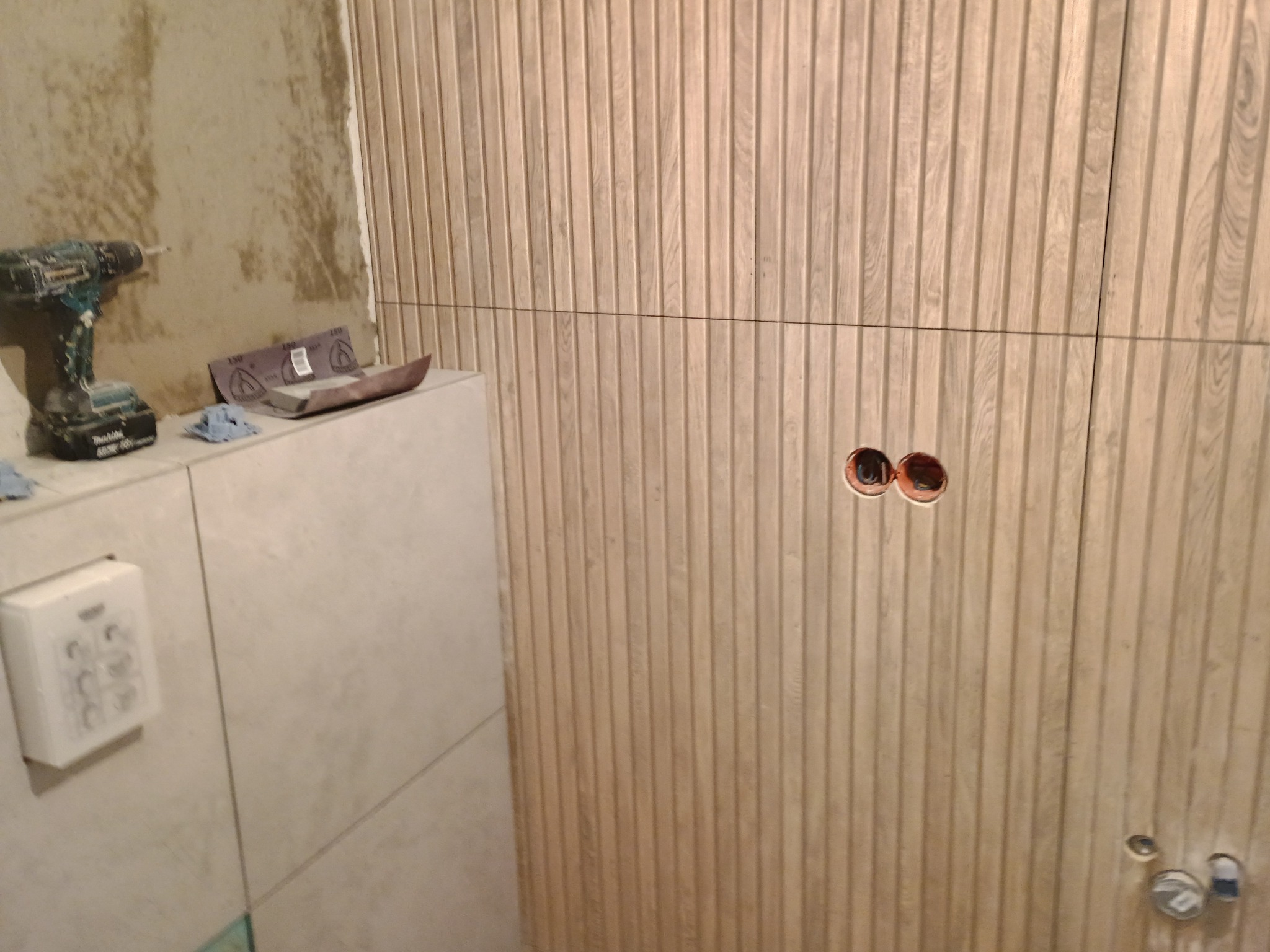 Łazienka w trakcie wykańczania: ściana z drewnopodobnych paneli z wyciętymi otworami na gniazdka, na blacie leży wiertarka akumulatorowa i papier ścierny.