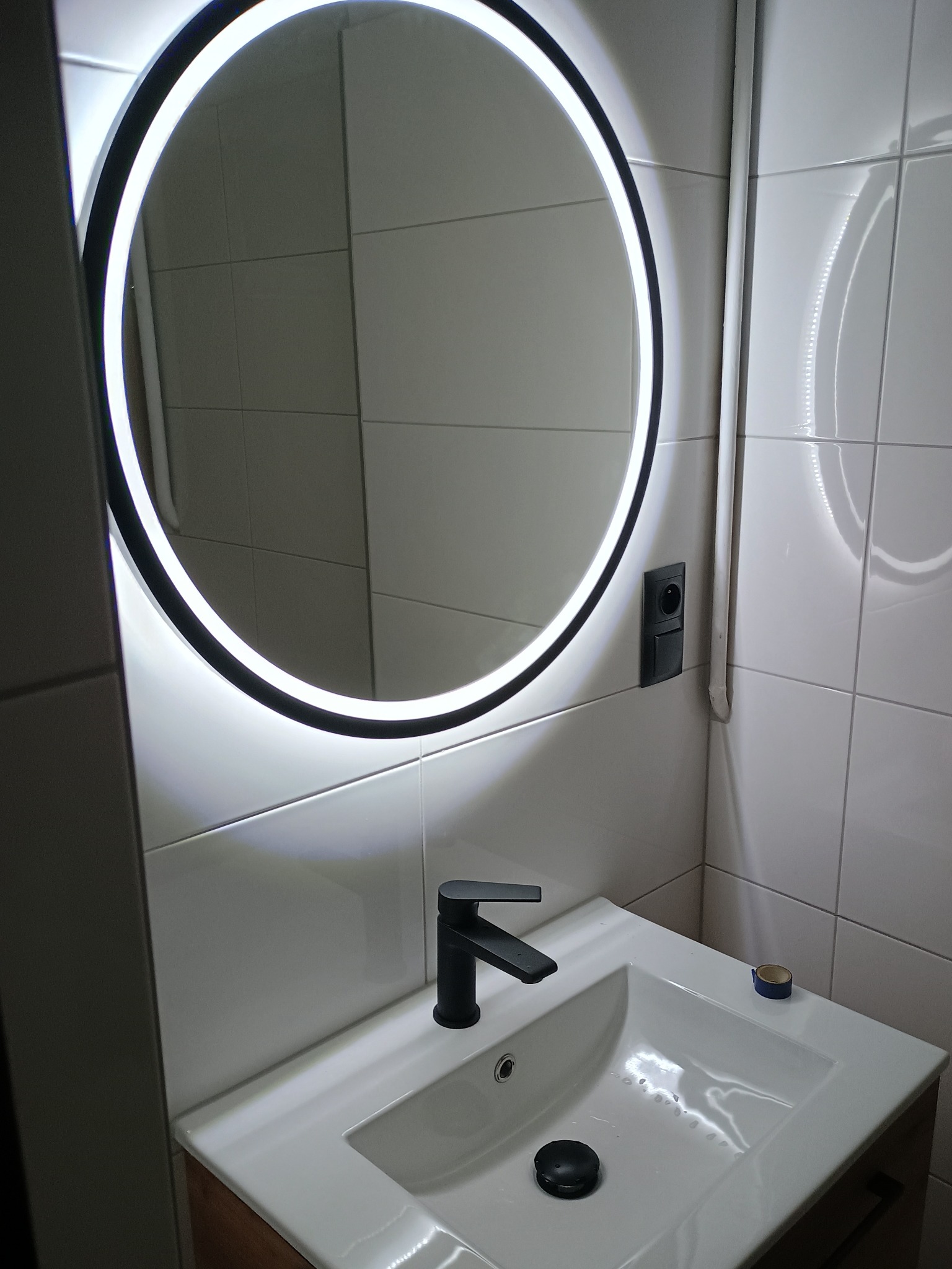 Nowoczesna łazienka z okrągłym lustrem LED, czarną armaturą i białą umywalką nablatową. Na blacie widoczny fragment niebieskiej taśmy malarskiej.