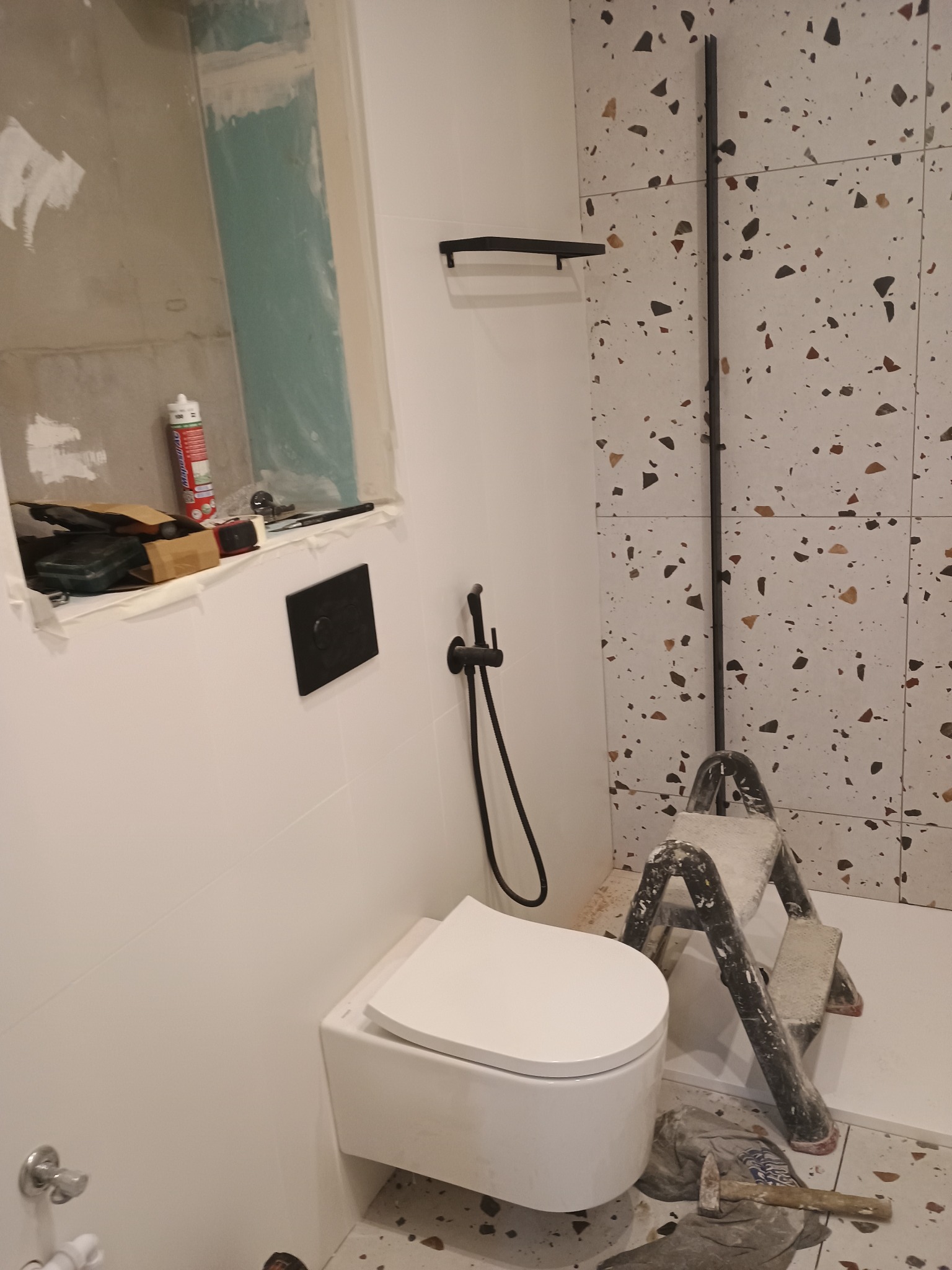 Remont łazienki w Strzyżowie: podwieszana toaleta, czarna armatura, płytki lastryko na ścianie, drabina malarska i narzędzia w trakcie montażu.