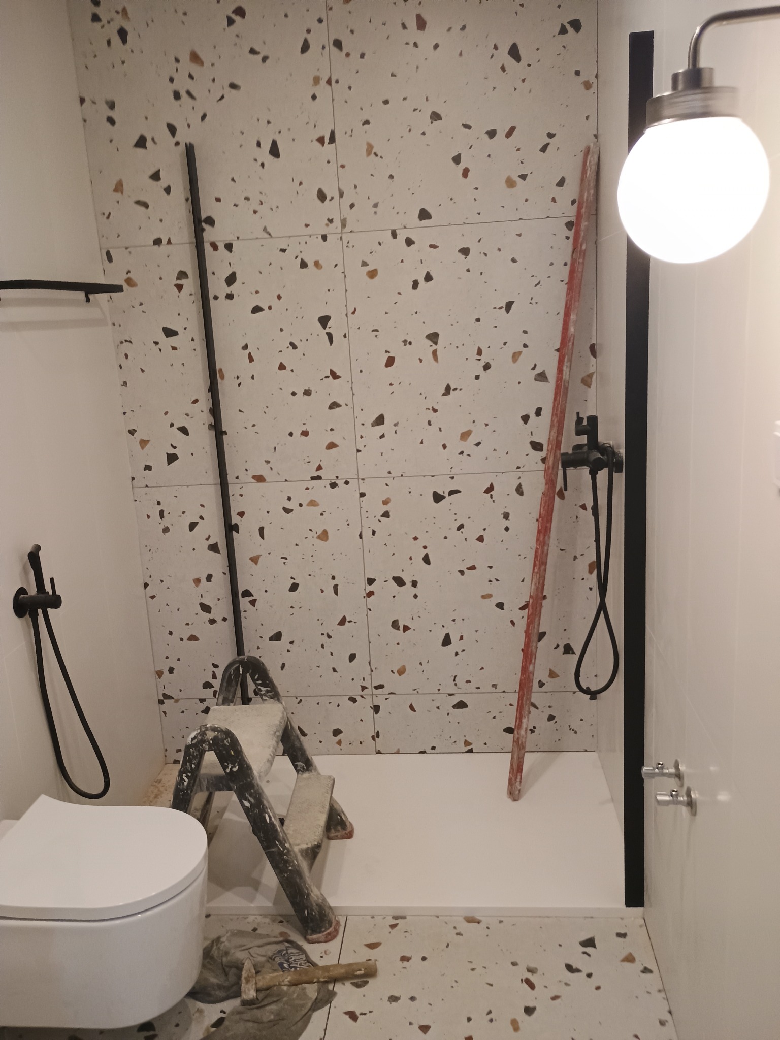 Nierównomiernie ułożone płytki lastryko na ścianie łazienki w trakcie prac wykończeniowych, widoczna drabina malarska, toaleta i czarna armatura prysznicowa.