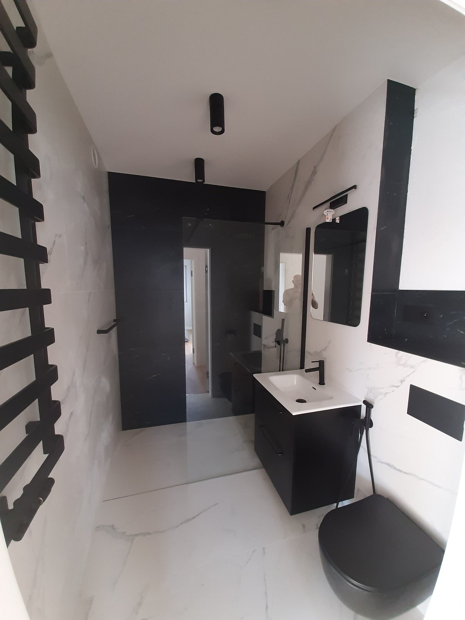 Nowoczesna łazienka z czarno-białymi płytkami imitującymi marmur, czarną armaturą i grzejnikiem drabinkowym oraz kabiną prysznicową typu walk-in z czarnymi elementami.