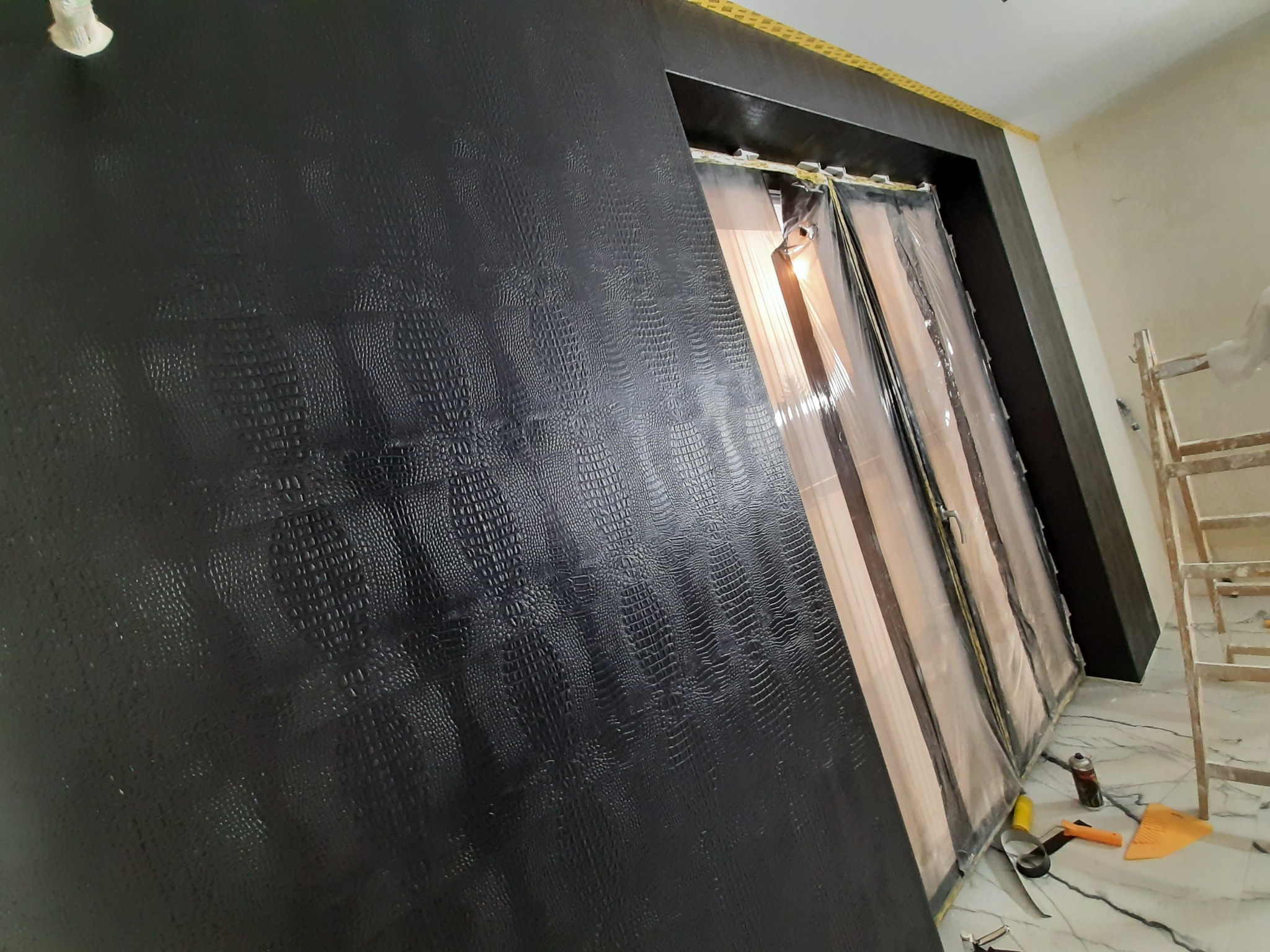 Ściana pomalowana na czarno z fakturą imitującą skórę krokodyla, zabezpieczone folią drzwi, drabina malarska i narzędzia na marmurowej podłodze w trakcie remontu.