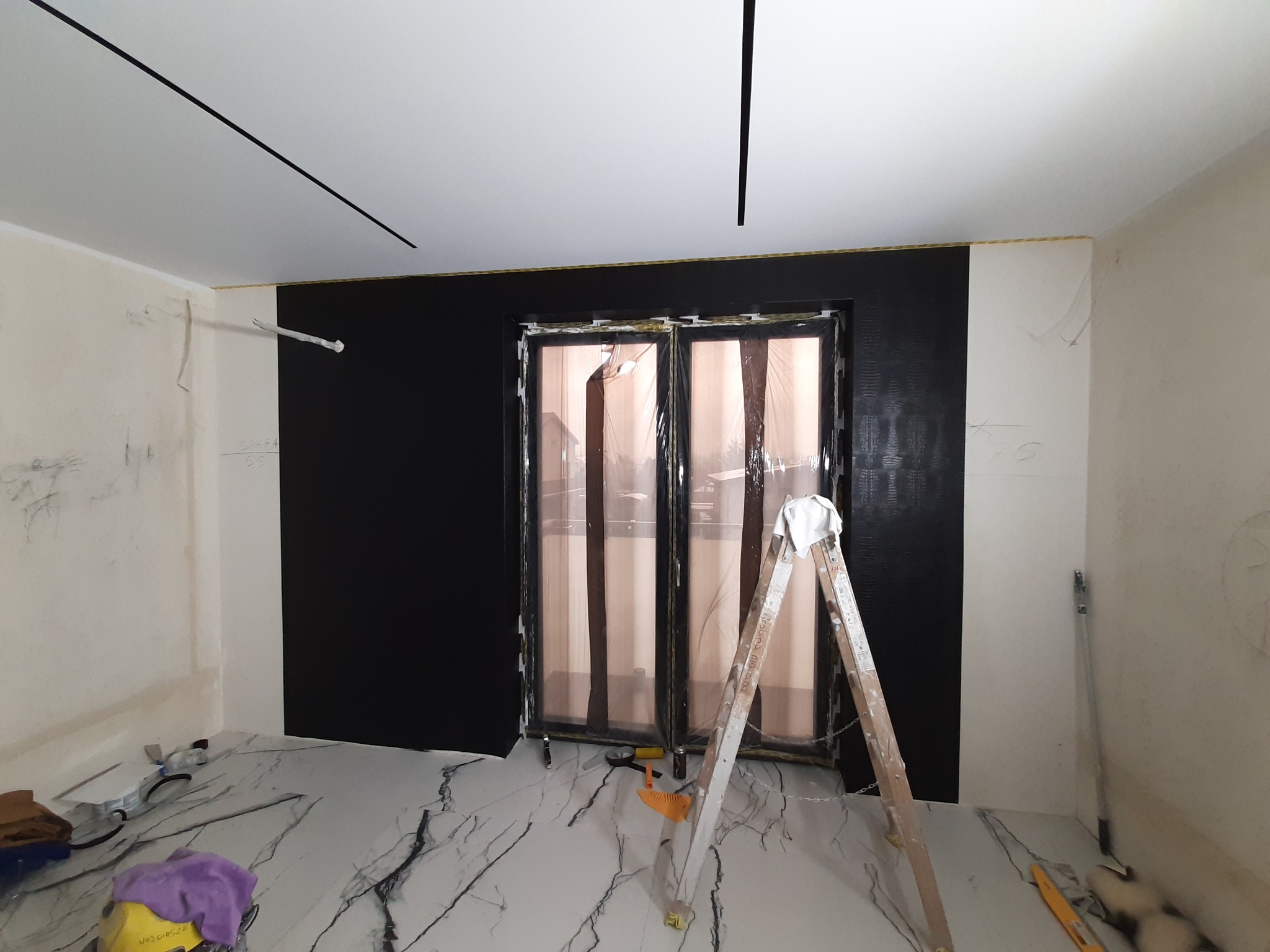 Pokój w trakcie malowania: ściana pomalowana na czarno, okno zabezpieczone folią, drabina oparta o ścianę, widoczne narzędzia malarskie na podłodze z marmurowym wzorem, sufit z czarnymi listwami.