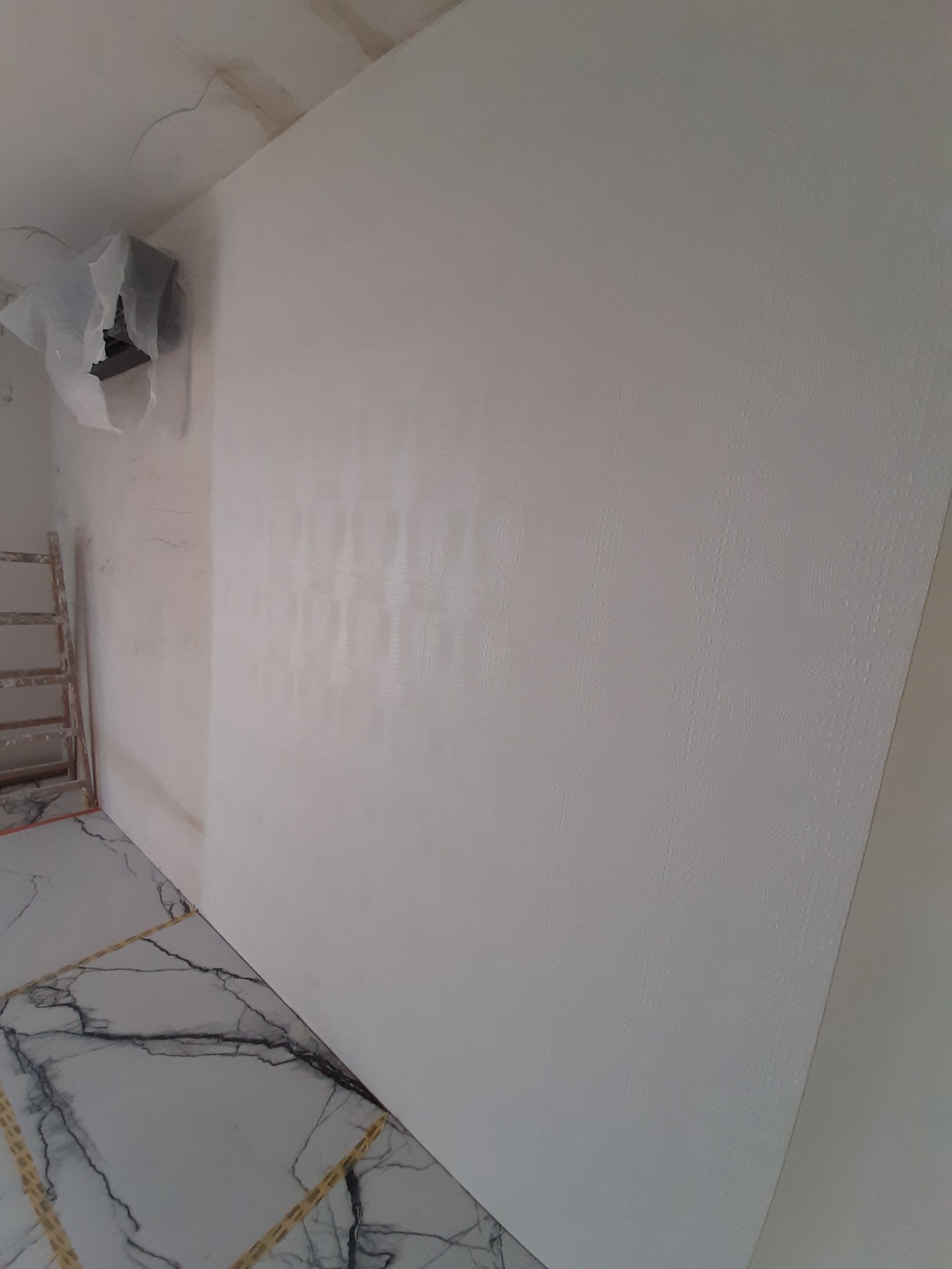 Świeżo pomalowana, gładka, biała ściana z delikatną fakturą, częściowo zakryty grzejnik, marmurowe płytki podłogowe z wyraźnym żyłowaniem, ustawiona drabina malarska