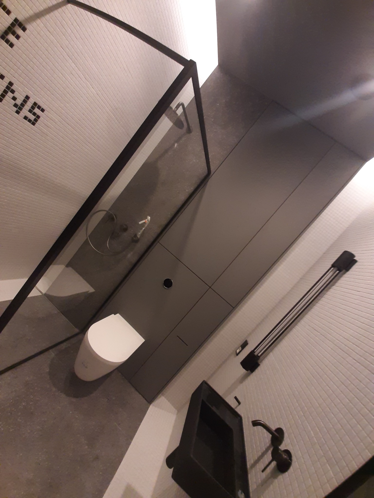 Nowoczesna łazienka z kabiną prysznicową typu walk-in z czarną ramą, podwieszana toaleta, szara zabudowa meblowa i białe płytki z czarnymi napisami.