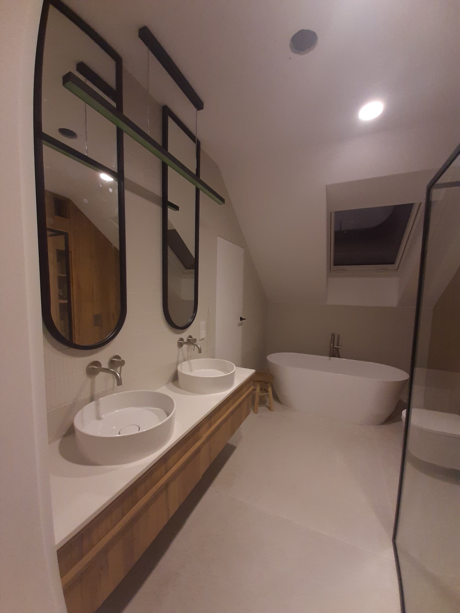 Nowoczesna łazienka na poddaszu z dwoma umywalkami nablatowymi, owalną wanną wolnostojącą i oknem dachowym; lustra w czarnych, podłużnych ramach z zielonym akcentem.