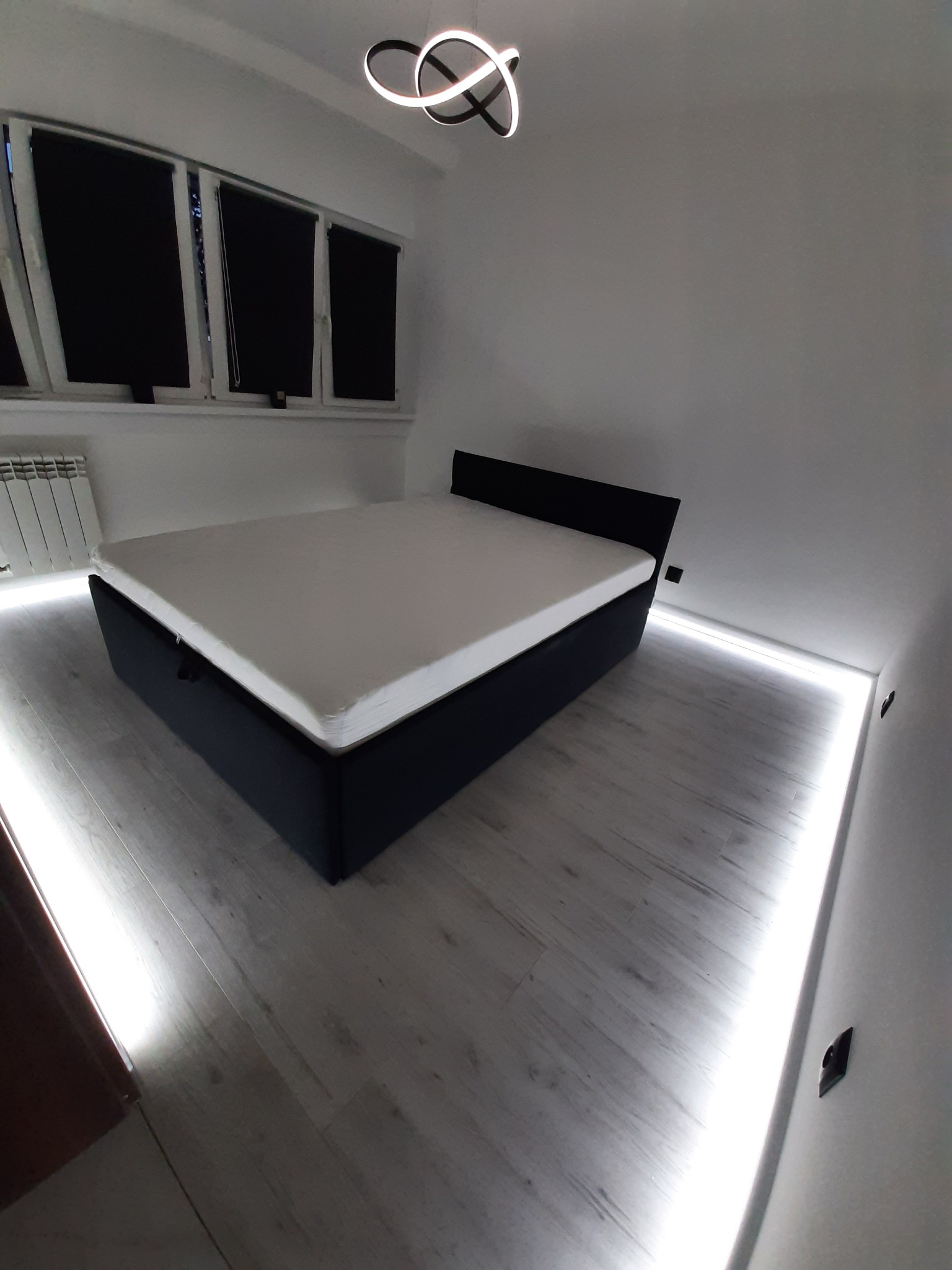 Nowoczesna sypialnia z łóżkiem kontynentalnym, oświetleniem LED pod sufitem i oknami z roletami w kolorze czarnym.