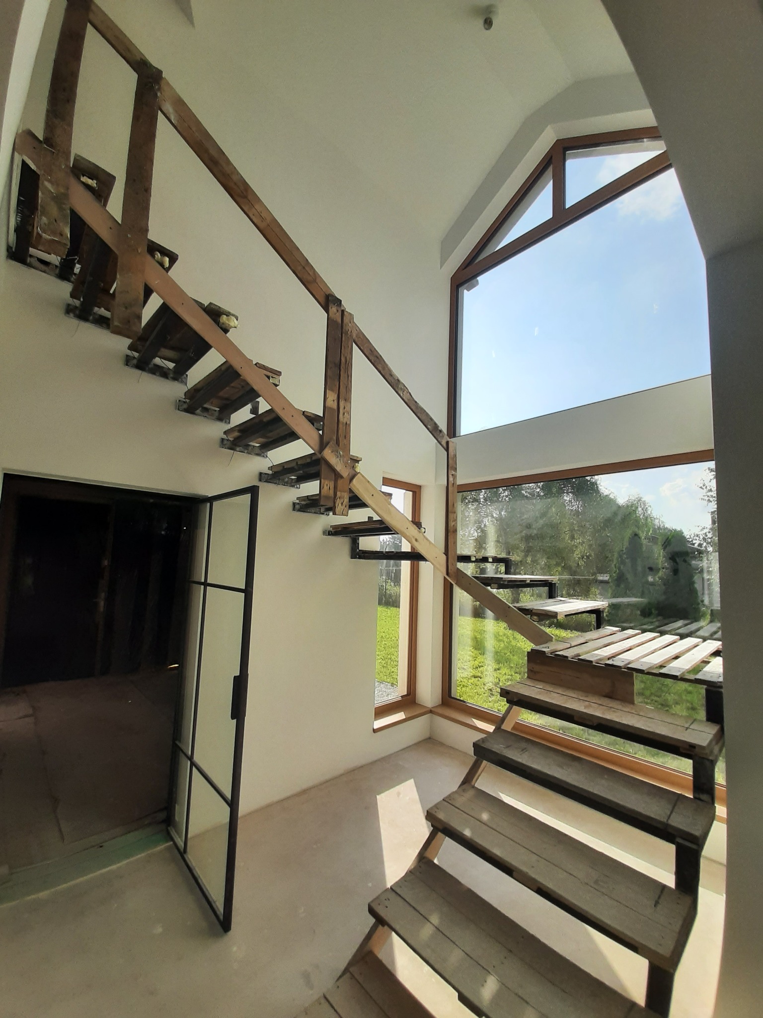 Wnętrze w trakcie remontu z drewnianymi schodami w surowym stanie, dużym oknem z widokiem na zieleń i otwartymi drzwiami do ciemnego pomieszczenia.