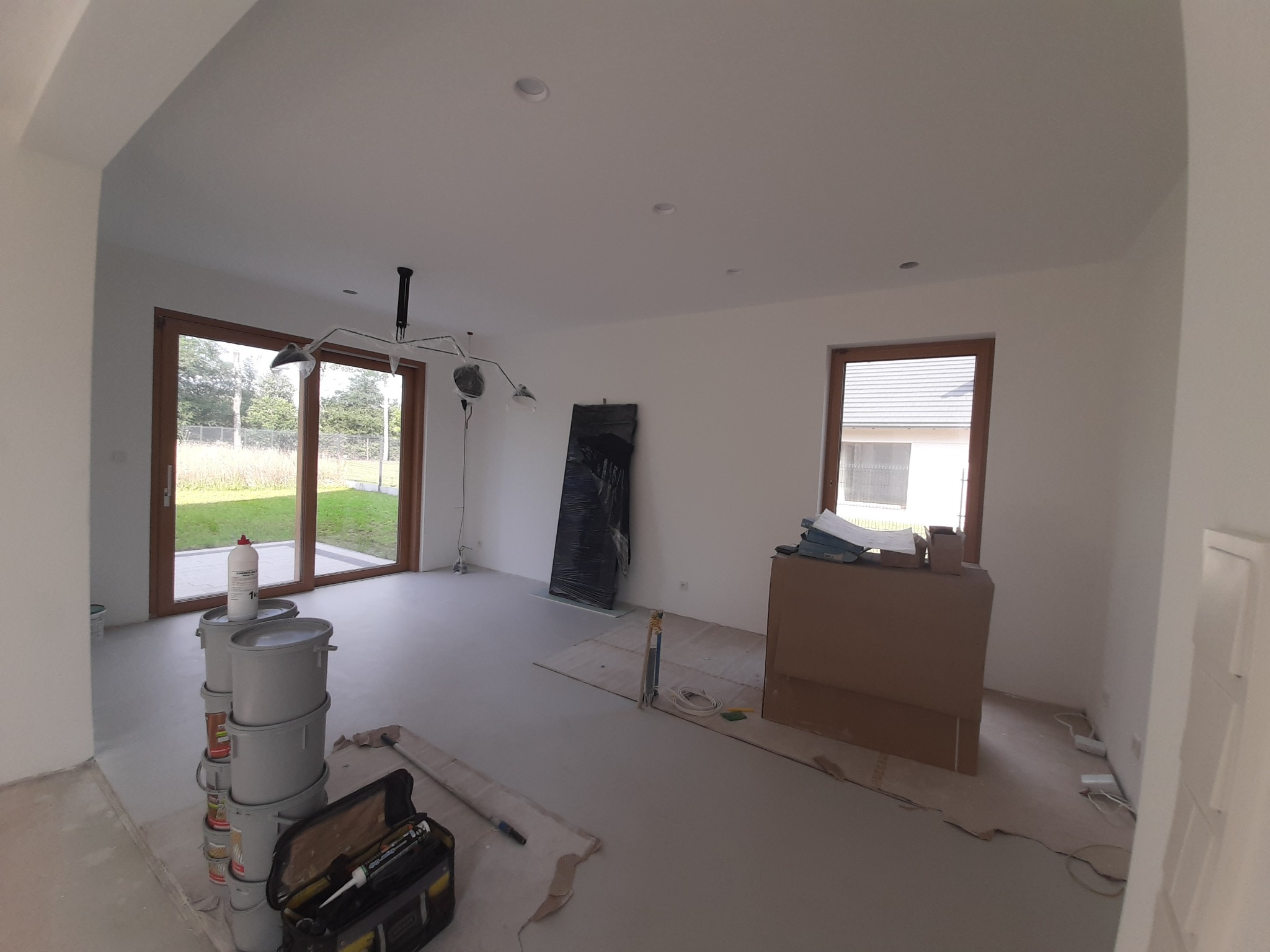 Wnętrze w trakcie malowania: widoczne puste ściany, zakryte folią drzwi, okna z drewnianymi ramami, puszki z farbą i narzędzia malarskie w otwartej skrzynce.