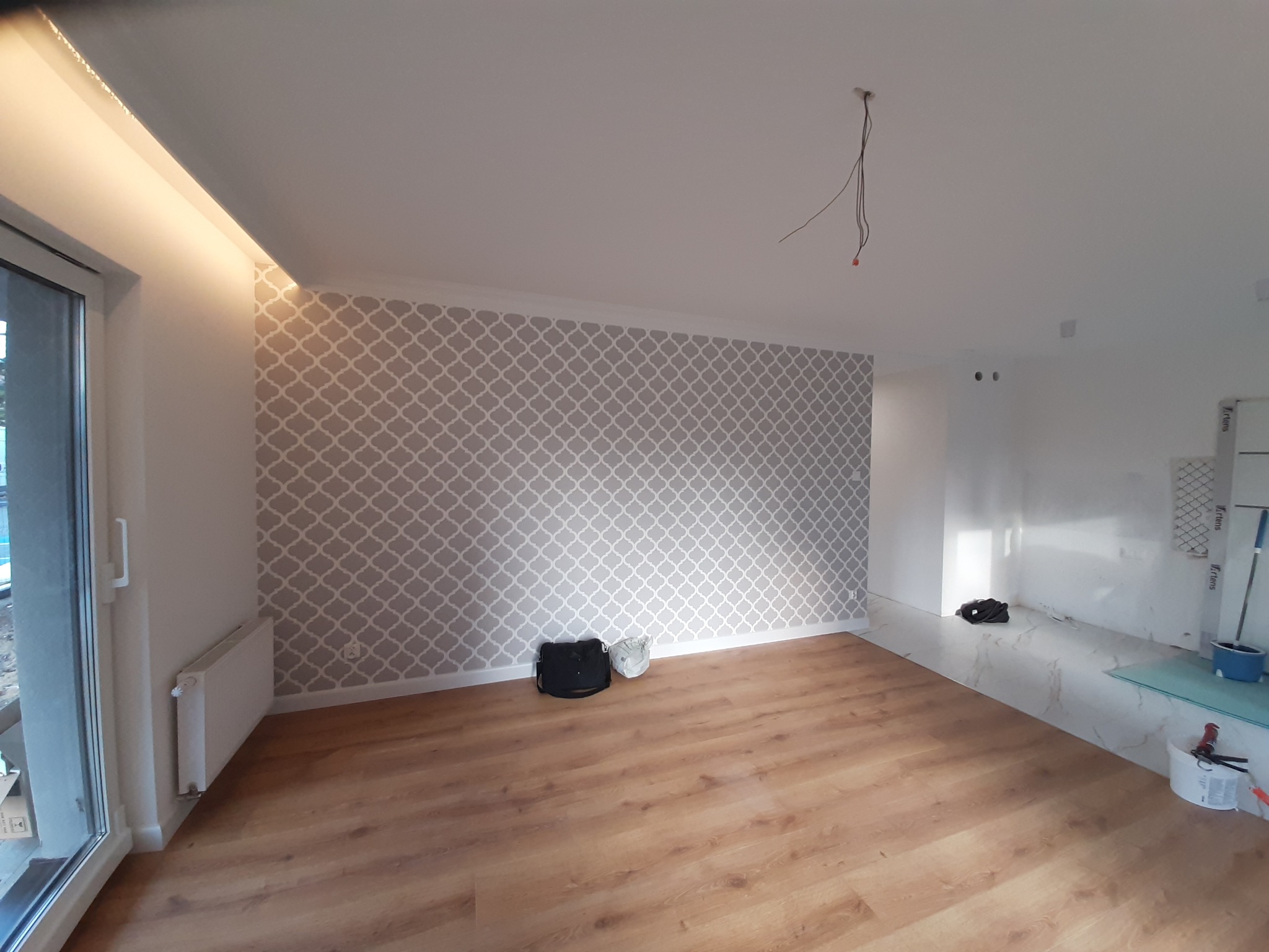 Wnętrze pokoju po remoncie, ściana z szarym wzorem, drewniana podłoga, listwa LED pod sufitem, widoczne kable wystające z sufitu.