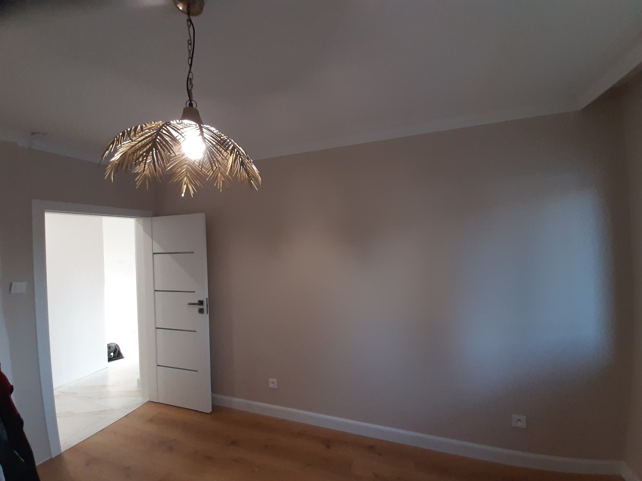 Pomalowane na beżowo wnętrze pokoju z drewnianą podłogą, widoczna biała, nowoczesna lampa w stylu boho oraz białe drzwi z poziomymi frezami.