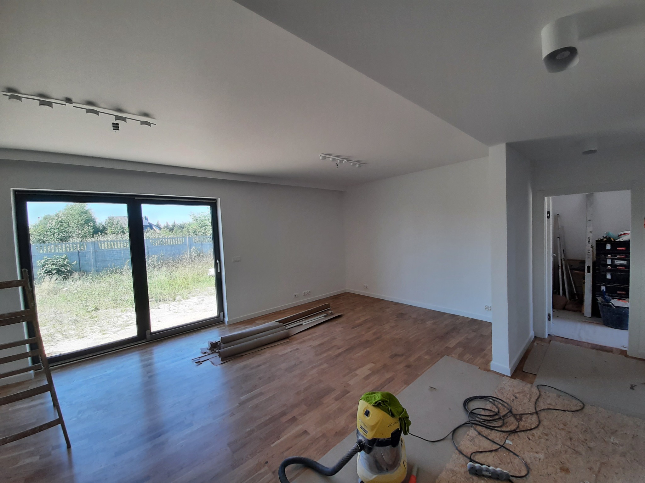 Jasne, przestronne wnętrze w trakcie remontu: świeżo pomalowane białe ściany, parkiet na podłodze, widoczne listwy przypodłogowe, drabina oparta o okno tarasowe z widokiem na zarośnięty ogród...
