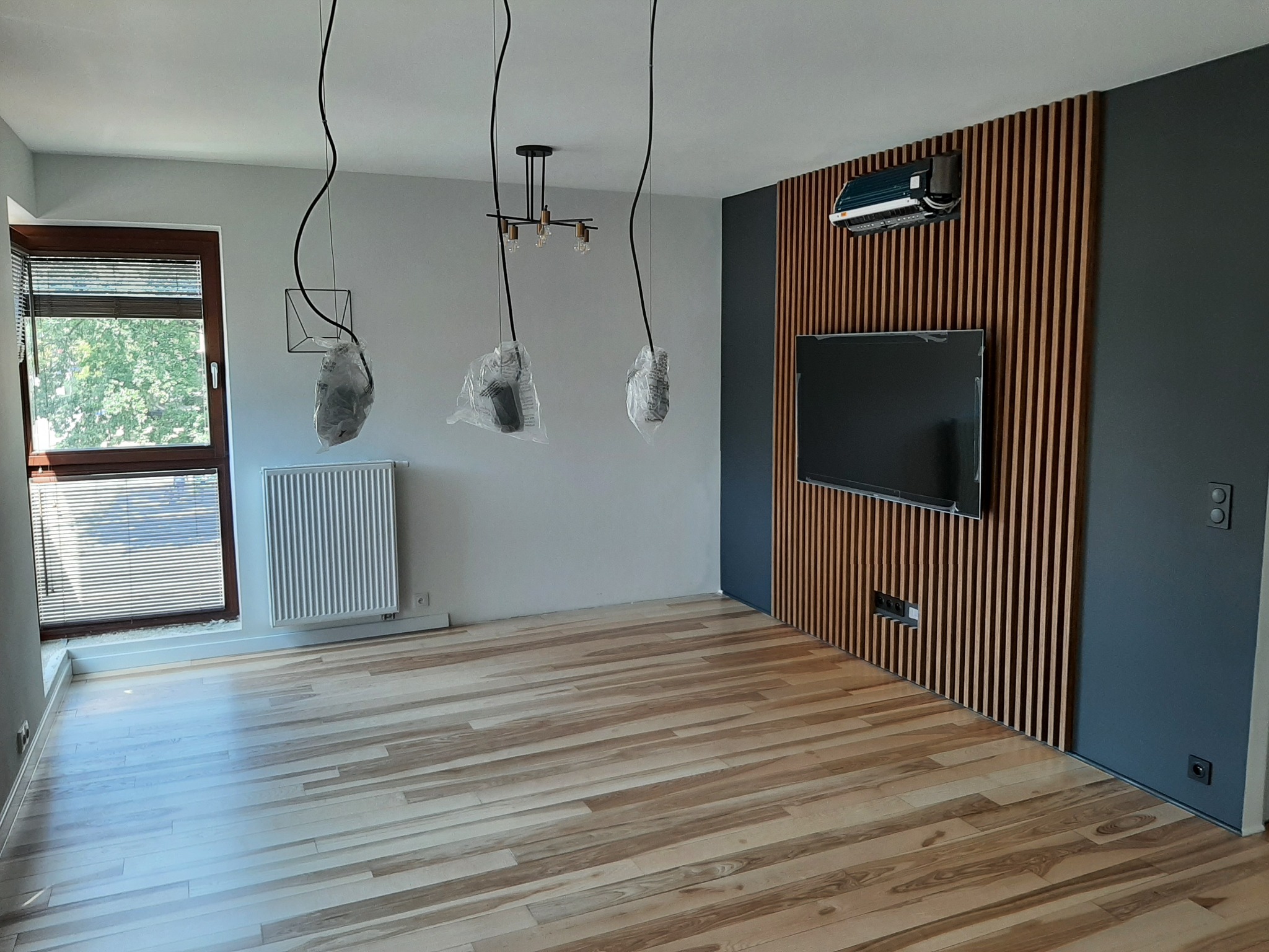 Wnętrze pokoju po remoncie z drewnianą ścianą z lamelami, klimatyzatorem, telewizorem, nową podłogą i lampami wiszącymi w folii ochronnej.