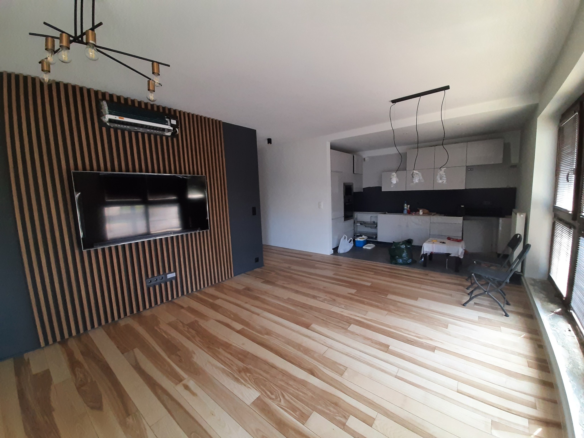 Nowoczesne wnętrze po remoncie: drewniana ściana z telewizorem, parkiet w jodełkę, kuchnia w tle z minimalistycznym oświetleniem, widoczny klimatyzator.