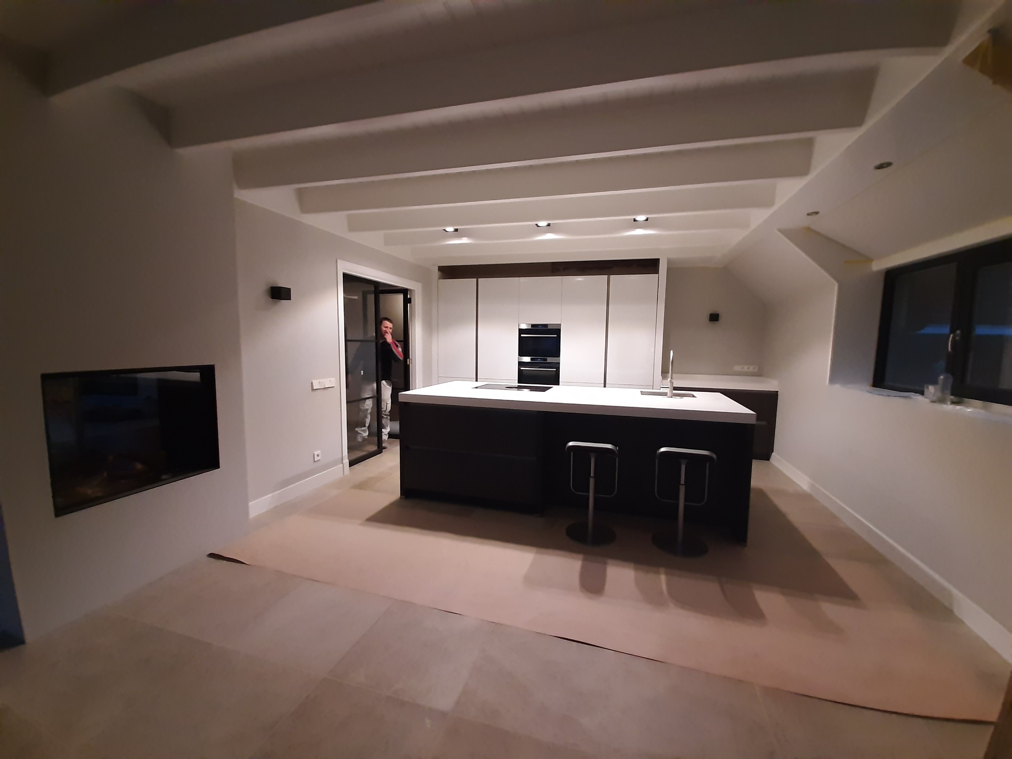Nowoczesna, jasna kuchnia z wyspą i dwoma hokerami. Podłoga zabezpieczona kartonem, widoczne oświetlenie sufitowe, minimalistyczne białe szafki i czarne akcenty. W tle widoczna osoba w drzwiach.
