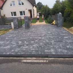 Judis SP. z o.o - Szeroki podjazd z szarej kostki brukowej, zakończony czarnym obrzeżem, prowadzący do domu z szarą, nowoczesną bramą w tle, widoczni ludzie.