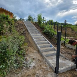 Judis SP. z o.o - Budowa schodów z kostki brukowej prowadzących na wzniesienie, w Nowogródku Pomorskim. Widoczny fragment ogrodzenia panelowego w tle i pracownik w trakcie montażu.