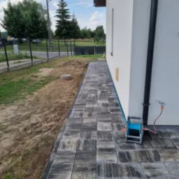 Ułożona ścieżka z szarej kostki brukowej obok białego domu. Widoczny trawnik z ogrodzeniem w tle i zwinięty wąż ogrodowy przy ścianie.