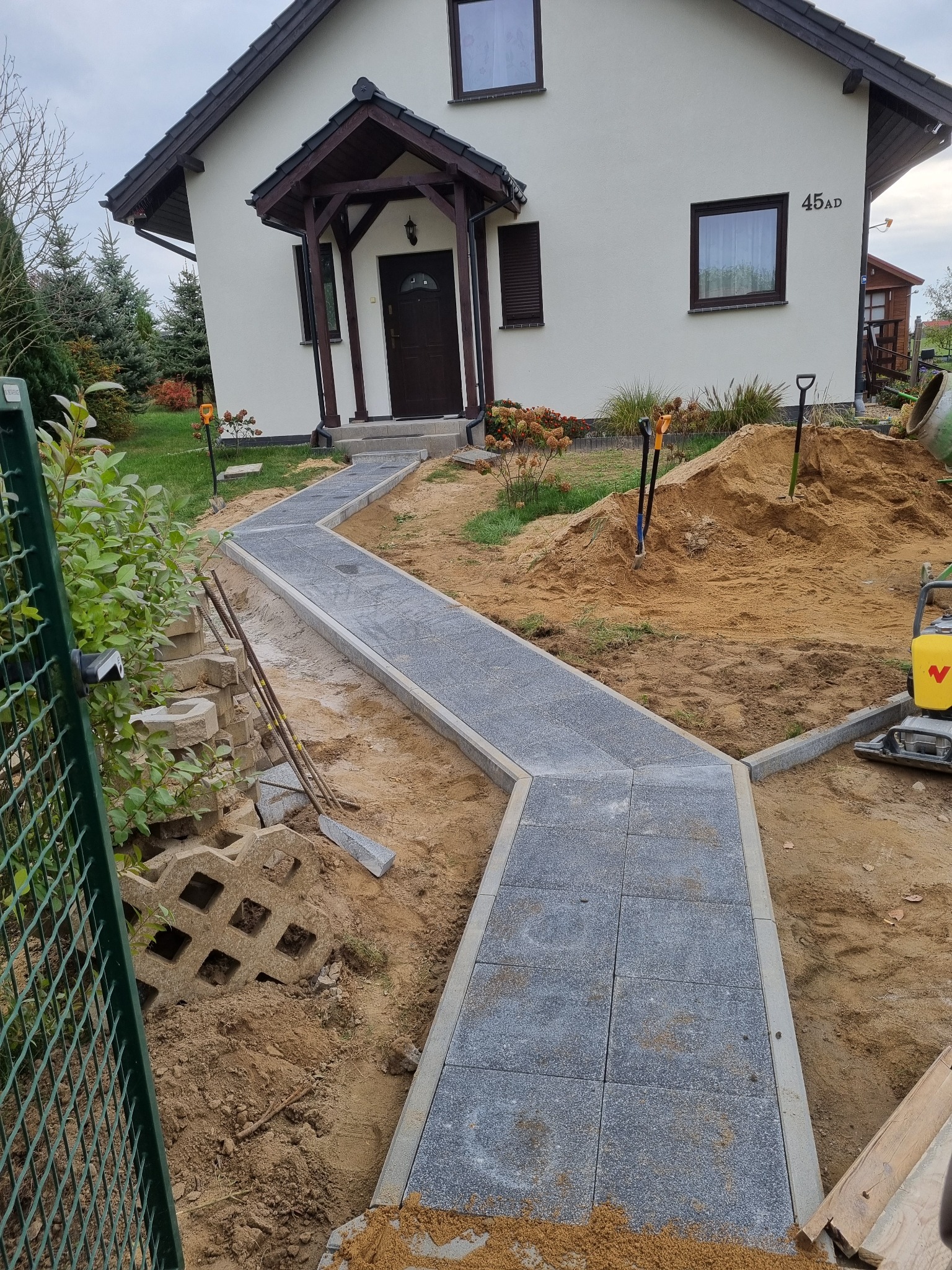 Ścieżka z szarej kostki prowadząca do domu, w trakcie budowy, widoczny ubijak wibracyjny, sterta piasku i narzędzia.