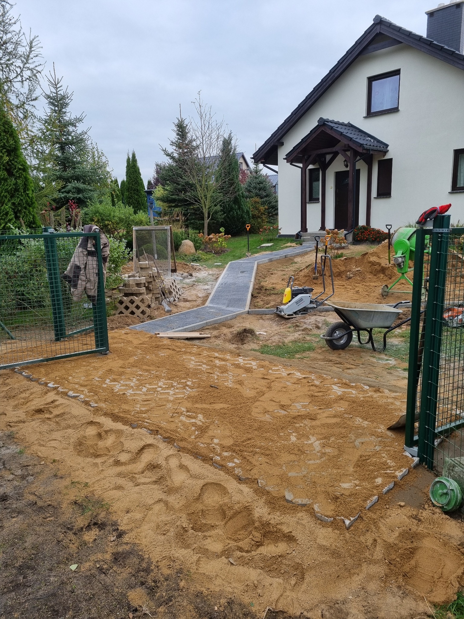 Trwa układanie ścieżki z kostki brukowej na posesji; widoczny fragment ułożonej ścieżki, ubijarka, taczka, betoniarka oraz przygotowany teren pod dalsze prace.