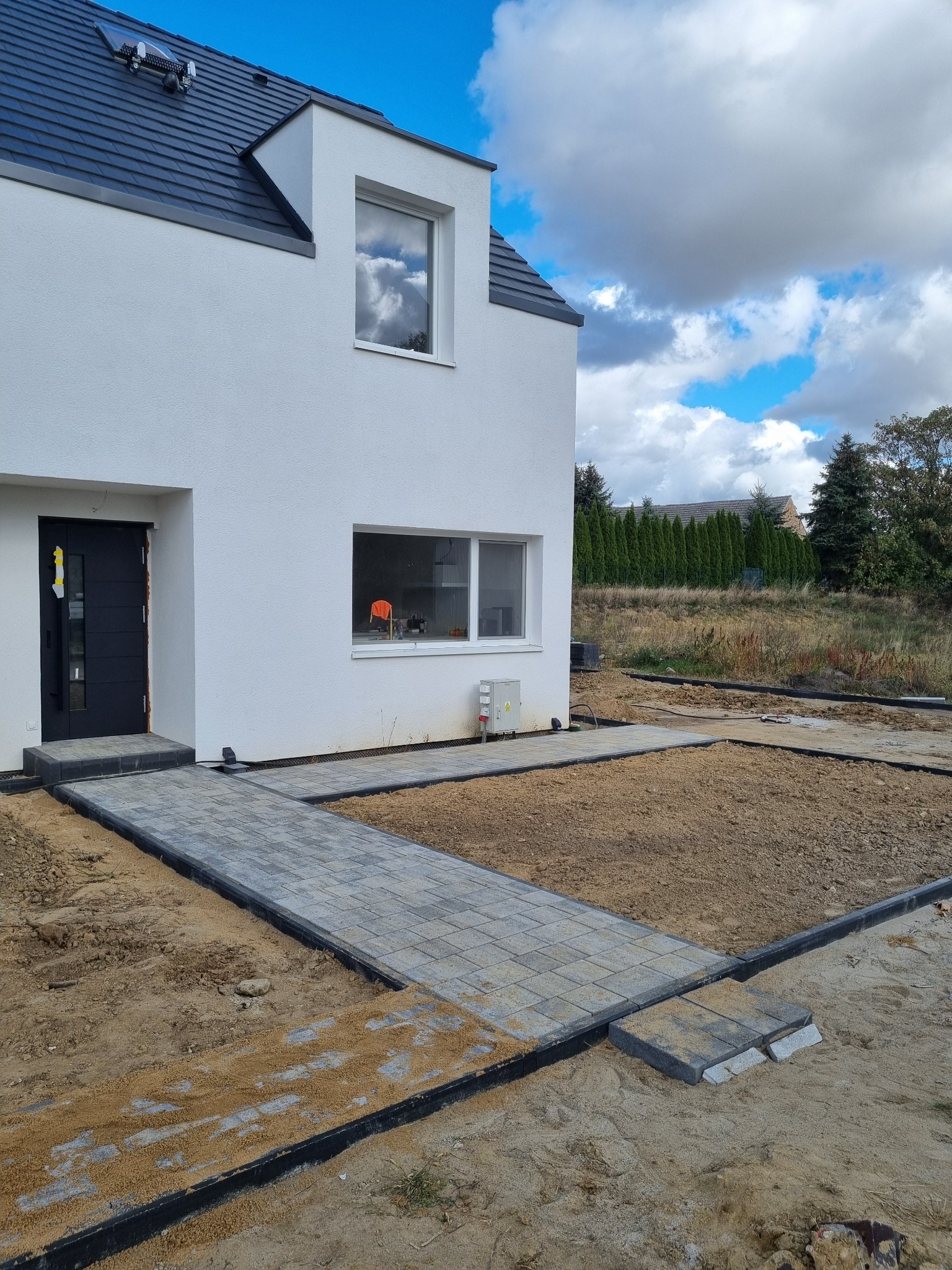 Nowoczesny dom jednorodzinny z wybrukowanym chodnikiem z szarej kostki, widok na ogród i błękitne niebo z chmurami.